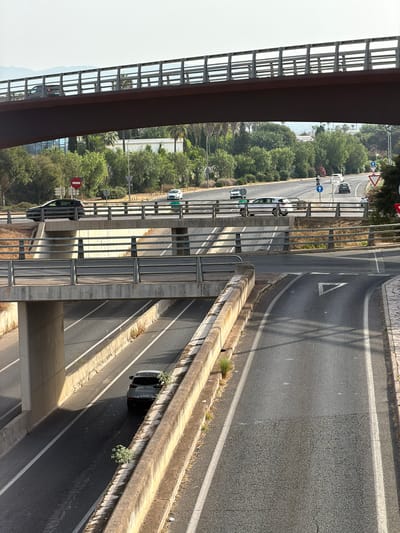 The image depicts an elevated highway interchange in Son Sardina, Spain. A dark-colored SUV is observed on the lower level of the highway, traveling away from the viewer. The vehicle has a license plate that reads "7615 LCM". On an upper level of the highway, a white car is seen traveling to the left. Further in the background, several other vehicles are visible on multiple levels of the interchange, with the landscape beyond showing trees and a hazy sky. The infrastructure consists of concrete overpasses and guardrails, with a clear division of traffic lanes indicated by white lines. The scene appears to be a busy road junction under bright daylight conditions.