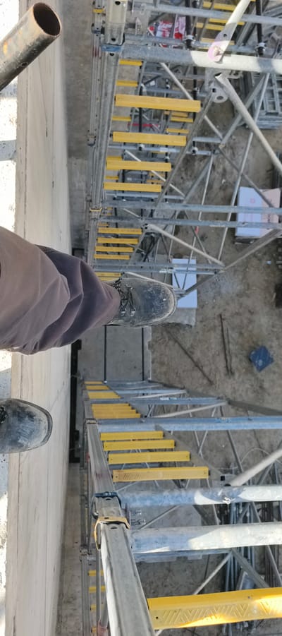 This image shows a first-person perspective of someone standing on scaffolding. The person is wearing work boots and dark pants. The scaffolding is made of metal and has yellow planks for steps. Below the scaffolding is dirt ground with some debris, including what appears to be construction materials. The scene suggests an outdoor construction site. No specific location in Primorye, Russia can be determined from the image alone.