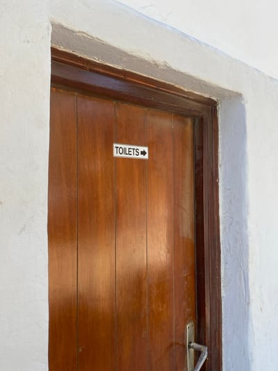 A close-up view captures a reddish-brown wooden door set within an off-white plastered wall. The scene is located in the city of Φάρος, Greece. The door features vertical paneling and a darker brown wooden frame. A white rectangular sign, approximately 10-15 cm in length, is affixed to the door's upper center, bearing the black text "TOILETS" followed by a black right-pointing arrow. A silver-colored metal door handle with a curved grip is present on the lower right portion of the door. The surrounding wall exhibits an uneven, textured finish consistent with aged plaster or stucco, with minor chips and surface irregularities visible along the door frame and upper edges. No individuals are present, and no actions are depicted within the static environment.