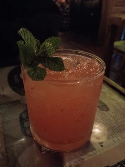 A close-up image of a pink drink in a clear glass, garnished with a sprig of mint. The drink is resting on a table that is covered in a brown and white patterned cloth.  The image was taken at latitude: -34.452, longitude: -58.54 in the city of Victoria, Argentina.  The drink is in focus and the background is blurred. The scene is inside, likely at a bar or restaurant.