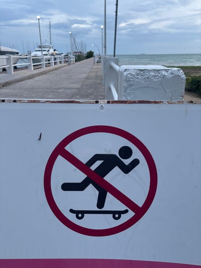 In Tambon Sattahip, Thailand, a "no skateboarding" sign is prominently displayed in the foreground. The sign features a black silhouette of a person on a skateboard, crossed out by a red circle and diagonal line. The sign is mounted on a white surface that appears to be weathered. In the background, a pier or walkway extends into the distance, lined with white railings and lampposts. Several boats, including large yachts, are docked along the pier, suggesting a marina or harbor setting. The sea and a cloudy sky form the backdrop, indicating an overcast day. The overall scene depicts a public area near the water, with regulations in place to manage recreational activities.