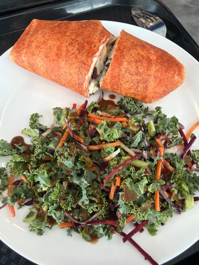 The image displays an overhead view of a meal served on a white plate, resting on a black tray, presumably in Mirabel, Canada. The primary subjects are a large, reddish-orange wrap and a side salad. The wrap is cut into two segments, revealing a filling of shredded light-colored vegetables, darker elements, and a creamy white dressing. The salad consists of dark green, curly kale leaves, julienned orange carrots, shredded purple cabbage, and thin white vegetable strips, all covered in a glossy, brown dressing. A silver fork with a textured handle is partially visible on the black tray in the upper right background. No people, actions, or interactions are visible.