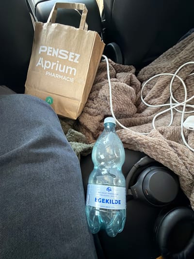 The image, taken inside a vehicle in Ödsmål, Sweden, shows a brown paper bag with the text "Pensez Aprium Pharmacie" printed in white. Next to the bag is a brown, fuzzy blanket. A clear plastic bottle of "EgeKilde" mineral water, labeled "UDEN BRUS / STILL," is positioned in front of the blanket. A pair of black Sony headphones rests on the blanket, and a white cable with an earbud is also visible. The lower left portion of the image displays a person's dark gray denim-clad leg. The scene appears to be a personal collection of items within a car.