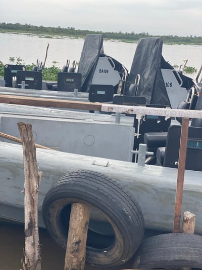 The image depicts a waterside scene at a rudimentary mooring point in Akarakumoh, Nigeria, under an overcast sky. In the foreground, several weathered wooden posts are partially submerged in dark water, supporting two black vehicle tires used as fenders against a boat hull. One tire is notably impaled by a wooden post. A horizontal wooden beam stretches across the lower mid-ground.
The mid-ground features multiple grey-painted patrol boats or rigid-hull inflatable boats (RHIBs) moored side-by-side. The most prominent boat displays the identifier "BA109" on its bow, which is partially covered by a dark grey tarpaulin. To its right, a similar boat with the identifier "106" is visible, also partially covered by a dark grey tarpaulin. Each visible boat is equipped with multiple black, individual seats and several black, rectangular armored panels featuring small, clear viewing slits positioned along the gunwales. Grey metal cleats are affixed to the boat hulls.
The background shows a wide, calm body of water, with patches of green aquatic vegetation, likely water hyacinths, floating near the distant bank. A dense line of green vegetation, consistent with trees or mangroves, forms the horizon under a uniform, light grey sky. No individuals are present in the scene.