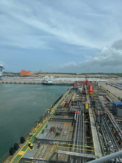 The image shows a view from a ship in a port, likely in Hambantota, Sri Lanka, looking towards the shore. The foreground features the deck of a large ship, characterized by numerous pipes, walkways, and industrial equipment. The ship's deck is a complex network of grey pipes of varying sizes, some marked with colored bands (red, blue, yellow). Yellow railings and ladders are visible, providing access to different levels. Black and yellow bollards are spaced along the deck.
The middle ground depicts a calm body of water, likely the harbor, separating the ship from the port facilities. A smaller blue and white boat is moored near the shore. Beyond the water, a long concrete pier is visible, lined with what appear to be parked cars, stretching across the frame. These vehicles are mostly white and densely packed, suggesting a car import or export terminal.
In the background, industrial buildings with orange and grey facades are prominent. Further inland, more structures, possibly storage facilities or a power plant, are discernible, along with masts and cranes typical of a port environment. The sky above is partly cloudy, with a mix of light and darker grey clouds against a pale blue sky. The overall scene suggests a bustling industrial port engaged in shipping and logistics, with a clear indication of car transport operations.