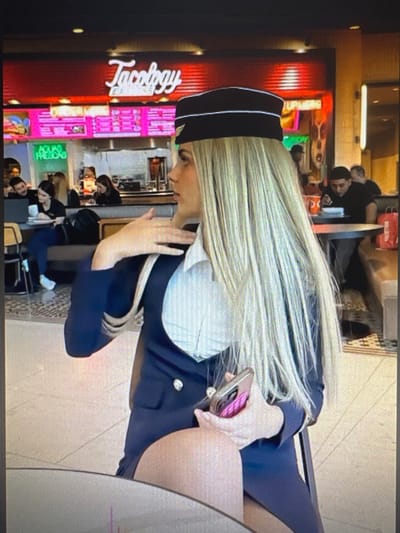 The image shows a person with long blonde hair wearing a dark blue uniform with a white collar and a black cap. The person is sitting at a table in what appears to be a food court or fast-food restaurant. They are holding a pink phone in their right hand. In the background, there are signs for "Tacobeyy" and other colorful advertisements, suggesting a commercial setting. Several other people are visible in the background, some of whom are also seated or standing. The lighting suggests it could be daytime. The scene is indoors, with a tiled floor visible. There is no clear indication of the weather or time of day beyond the indoor lighting. The focus is on the person in the uniform.