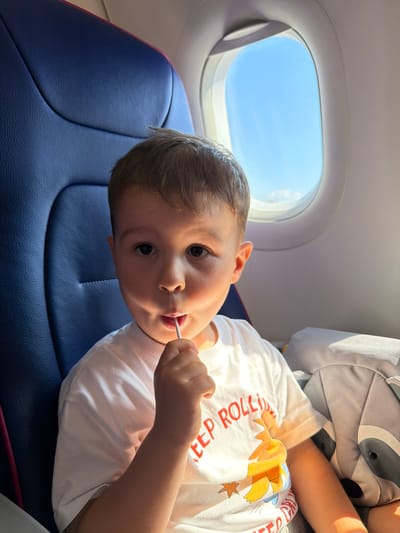 A young boy sits in an airplane seat, holding a lollipop to his mouth. He is wearing a white t-shirt with a graphic design and red lettering. To his right, a grey backpack with a raccoon design is visible. Behind him, a blue airplane seat is present, and to his left, an airplane window shows a bright blue sky with scattered clouds. The scene appears to be within the cabin of a commercial aircraft, likely en route to or from Donja Cijevna, Montenegro, given the context provided.