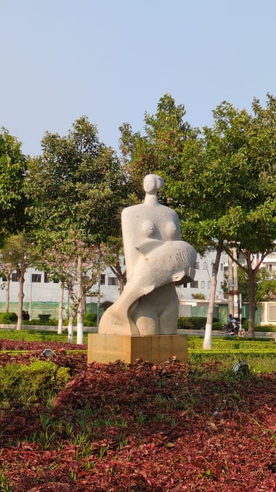 The image features a marble sculpture of a reclining woman with a curved, smooth form placed on a stone pedestal, situated in a park setting. The sculpture sits amidst lush greenery, including trees and what appears to be a dense bed of red-leaved ground cover. In the background, there are white buildings with windows and a glimpse of what may be a street with utility poles and possibly a motorbike. The sky is light blue, suggesting either early morning or late afternoon light. The location is indicated as being in the city of Phan Rang – Tháp Chàm, Vietnam.