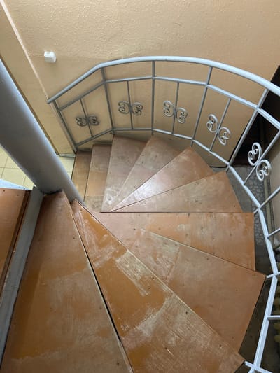 The image shows a spiral staircase viewed from above. The steps are made of wood and appear to be unfinished or worn, with visible scuff marks and a slightly rough texture. A gray metal pole runs along the inner edge of the staircase, serving as a central support. The railing along the outer edge is made of white metal with decorative scrollwork. The walls and ceiling are painted a pale yellow or beige color. There is a small white smoke detector mounted on the ceiling. The scene appears to be indoors, possibly in a residential building or a public space. The location is identified as Kraskino, Russia. There are no people or animals visible in the image.