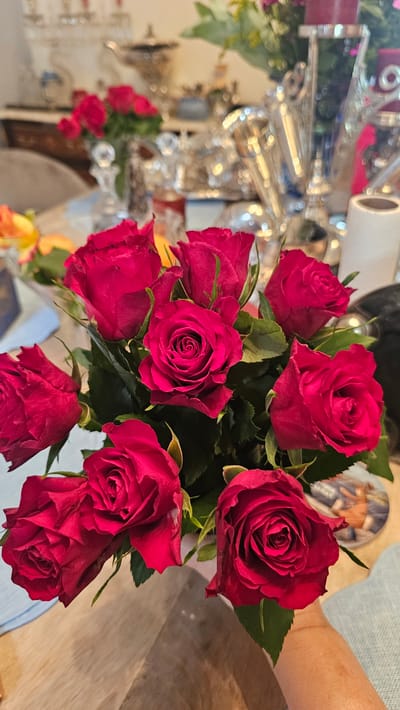 This is a close-up, eye-level shot of a bouquet of deep red roses in a vase, set on a wooden table. The roses are in various stages of bloom, with some tightly closed and others beginning to unfurl. The background is blurred, but appears to be an indoor setting with decorative silver objects and a vase of greenery. The lighting is soft and warm, creating a slightly romantic or celebratory atmosphere. There are no people visible in the frame. The image does not provide any information about the location in Charleroi, Belgium.
