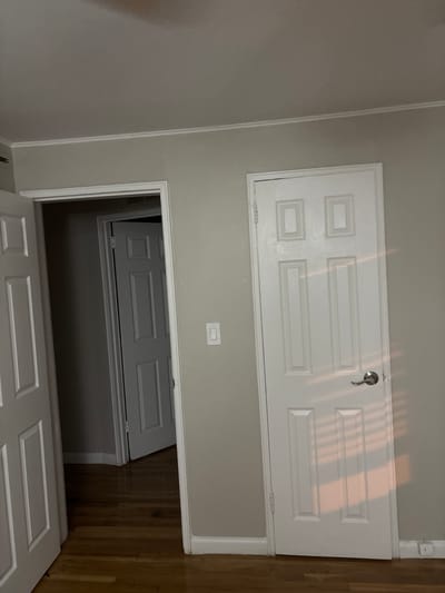 The image depicts an interior residential space, possibly a hallway or transitional area, with multiple doors and a wooden floor. The photograph is oriented vertically, showing a light-colored wall with white trim.
In the upper part of the frame, a white, paneled door is prominently featured. This door, which appears to be a smaller utility access door or an attic door, is closed and has a silver-toned, lever-style handle on its right side. The door's design includes two smaller rectangular panels on the left and two larger rectangular panels on the right. Sunlight or artificial light casts distinct striped reflections diagonally across the surface of this door, suggesting it is daytime.
Below this upper door, on the wall, there is a rectangular white light switch. Further down, there is an open doorway leading into another section of the residence. Within this open doorway, a third white, paneled door is partially visible. This third door appears to be slightly ajar and leads into a darker space beyond.
The walls are painted a uniform, light grey or beige color. To the right side of the lower doorway, a light brown hardwood floor is visible, extending into the foreground. No people or visible text are present in the image.