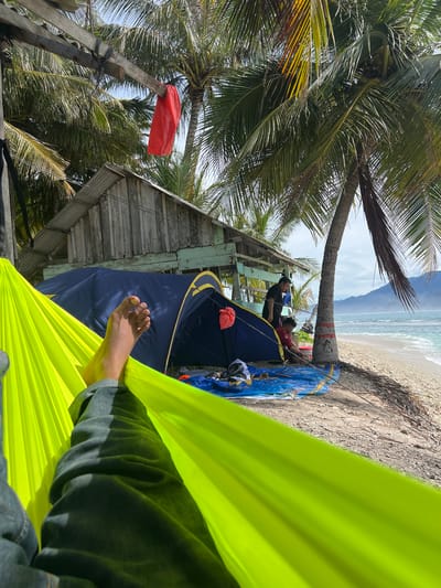 This is a first-person perspective shot from a bright green hammock on a tropical beach. In the background, a rustic wooden shack with a corrugated metal roof sits near a blue tent on the sand. Palm trees with lush green fronds tower over the scene, some showing evidence of coconuts. Two men are visible near the tent; one standing and wearing a dark t-shirt, the other, a young boy in a red shirt, is sitting. A red flag hangs from a wooden beam above the shack. The ocean waves gently break on the pebble-strewn shore, and in the distance, misty mountains are visible. The lighting suggests it is daytime with a clear sky. The location is identified as Babahie, Indonesia.