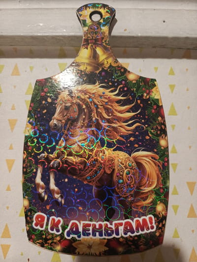 The image shows a decorative cutting board with a holographic design. The main subject is a majestic, ornate horse, adorned with golden armor and jewels, captured mid-stride with its front legs raised. The horse has a flowing golden mane and tail, and its head is turned to the left. The background is a dark, starry sky sprinkled with confetti or glitter. Surrounding the horse is a festive, Christmas-themed border featuring holly branches, red berries, golden baubles, and pinecones. At the top of the cutting board, there are two large golden bells tied with a red ribbon, and a small circular hole for hanging. At the bottom, there is text in large, bubbly, holographic letters. The overall aesthetic is celebratory and extravagant, likely intended for the New Year holiday, as indicated by the Christmas elements and the celebratory design. The text reads "Я к ДЕНЬГАМ!" which translates to "I am for money!" or "Towards money!". The lighting in the image suggests an indoor setting, possibly a home, with the holographic effect reflecting light and creating rainbow-like patterns on the design. The background surface appears to be a patterned tablecloth or placemat with a geometric design of yellow triangles.
