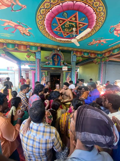 The image depicts the interior of a Hindu temple in Chinnambedu, India. The ceiling is elaborately painted with religious motifs and figures against a teal background. A white ceiling fan is visible. The walls and pillars are also decorated with vibrant colors and patterns. In the foreground, a crowd of people, both men and women, are gathered. Many are looking towards the center of the temple where a deity is likely enshrined. Some individuals appear to be engaged in prayer or religious rituals. The attire of the people suggests a religious ceremony or gathering. The overall atmosphere appears to be one of devotion and community.