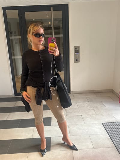 An individual stands in an indoor hallway or lobby of a building in Nogent-sur-Marne, France. The subject is a female with blonde hair, wearing black sunglasses, a black long-sleeved button-front top, and light beige ribbed capri-length pants. A black garment is tied around her waist. She wears black pointed-toe kitten-heel mules with stud embellishments. On her right hand, she holds a purple smartphone with a yellow pop-it case, displaying her reflection. White wired earbuds are connected to the phone. Multiple bracelets adorn her left wrist. The background features dark-framed glass double doors on the left, reflecting the reverse text "RDC" (Rez-de-chaussée). A light-colored wall on the right hosts a grey access control panel with a digital display and keypad. The floor is covered with light and dark grey rectangular tiles, and a dark grey ribbed mat is visible on the far right.