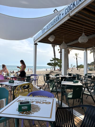 The image depicts an outdoor dining area at a beachside establishment identified by the signage "BAR RESTAURANT COCKTAILS" and "GOLDEN BEACH CLUB" visible on table covers and a banner. The scene is set in Cambrils, Spain. The foreground shows several tables and chairs on a wooden deck. One table in the center has a white surface with "GOLDEN BEACH CLUB" printed on it, along with a green box labeled "MAGNA CERVEZA RUBLA." A woman with dark hair is standing and gesturing with her left hand while wearing a lime green top and a black skirt. Another woman with blonde hair in a bun is seated at a table to her left, wearing glasses and a white top. In the background, a sandy beach extends to the sea under a blue sky. People are visible on the beach, some sunbathing and others walking. Palm trees and other vegetation line the edge of the beach, and a white canopy stretches over the dining area. The overall atmosphere suggests a relaxed, seaside dining experience.