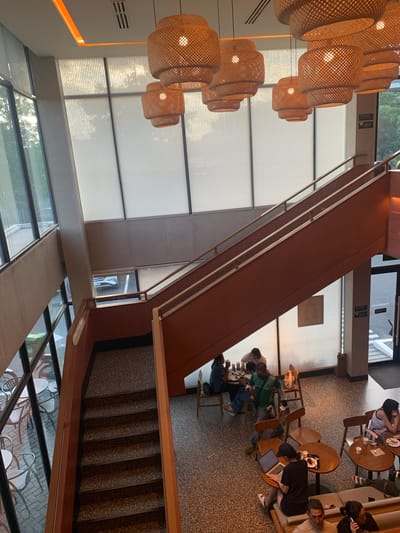 This is an indoor shot of a multi-level establishment, possibly a cafe or restaurant, in Angeles City, Philippines. The scene is captured from an elevated perspective, looking down on a staircase and an open area with seating.
Dominating the upper part of the image are several large, woven pendant lights, casting a warm glow. A grand staircase, with terracotta-colored risers and a wooden handrail, spans diagonally across the frame, leading to an upper level. The staircase has a speckled grey tread.
Below and to the right of the staircase, tables and chairs are arranged. Several people are seated at these tables, engaged in various activities. One person is working on a laptop, while others appear to be conversing and eating. A few individuals are standing or in motion, suggesting a dynamic environment.
The overall ambiance is modern and inviting, with large windows allowing natural light to filter in, creating a bright and airy atmosphere. The flooring is a speckled terrazzo, and the walls are a muted tone. To the right, a doorway with a glass panel is visible, possibly an entrance or exit.