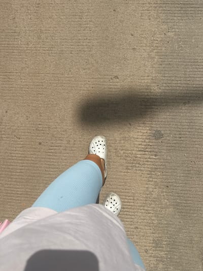 The image shows a first-person perspective of a person walking on a concrete surface. The concrete has parallel grooved lines running horizontally across the frame. The person is wearing white Croc-style shoes and light blue ribbed leggings. A portion of a white garment, possibly a t-shirt or dress, is visible in the foreground, obscuring part of the lower body. A shadow is cast on the concrete from an unseen object, likely from the sun. The image does not provide any specific visual cues to determine the location within the city of Xayden, Laos.
