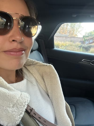 A person wearing sunglasses and a jacket with a cream-colored, fluffy lining is shown in a close-up, angled view. The person is positioned inside a vehicle. The car's interior is visible, with a window showing blurred scenery and greenery outside. The person is partially reclined, appearing relaxed or possibly sleeping. A small part of a seat and seatbelt are also visible in the background.