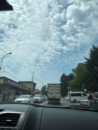 The image depicts a street scene in Novomikhaylovsky, Russia, viewed from the interior of a vehicle. The sky is filled with white, scattered clouds. In the foreground, the dashboard of a car is visible. The street is lined with shops and buildings on the left, displaying signage in Russian, including "ВСЕ ДЛЯ ДОМА" (Everything for the home) and "ОВОЩИ ФРУКТЫ ОДЕЖДА" (Vegetables, fruits, clothes). Various vehicles are present, including a white sedan, a white SUV, a white minivan, and a white truck with a shipping container. A woman wearing a black tank top and blue jeans is standing near a white car on the right side of the road. Trees are visible on the right side of the image.