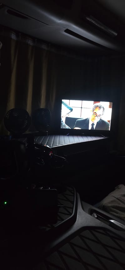 The image depicts an interior scene, likely a recreational vehicle or truck cab, with a television screen displaying a man in a suit and glasses. Two black electric fans are positioned to the left of the television. The environment is dimly lit, with beige curtains forming a backdrop behind the fans and television. A textured black surface, possibly a dashboard or seat, occupies the foreground. A small green light is visible on the lower left side of the frame. The overall impression is one of a private, possibly resting, space. There is no identifiable information in the image that would indicate the location is the city of Épila, Spain.