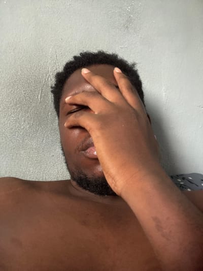 The image displays a male individual with dark skin, short dark hair, and a visible beard, situated indoors in Gbongan, Nigeria. His right hand covers his eyes and forehead. His left shoulder and upper chest are exposed, indicating he is shirtless or partially clothed. The background consists of a light grey textured wall, and a dark fabric with a light patterned design is partially visible behind his right shoulder on the right side of the frame.