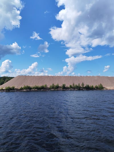 The image shows a large pile of light brown gravel or sand on the shore of a body of dark blue water. The gravel pile is massive, with a wavy, tiered surface, and is topped with a line of green trees and bushes. In the background, a dense forest of green trees covers a hill. The sky above is bright blue with scattered white, puffy clouds. The water in the foreground is choppy with small waves. A thin, light-colored pole extends from the water towards the gravel pile. The scene appears to be a natural landscape or a man-made industrial site, likely a quarry or a material storage area. The location is identified as Kvartsitny, Russia.