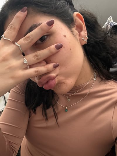 The image displays a close-up portrait of a female individual, located in AMCO, Area Metropolitana Centro Occidente, Colombia. Her right hand is positioned across the upper part of her face, partially obscuring the left side of her nose and mouth. Her fingernails are painted a muted purplish-brown color. She wears three silver-colored rings on her right hand: a wavy band on the ring finger, a ring with a black segment on the middle finger, and a small heart-shaped ring on the index finger. Her visible right eye is open and directed towards the viewer. Her lips are pursed in a pronounced pout, with a portion of her tongue visible. Several small moles are present on her right cheek and jawline. Her right ear is visible, featuring a small stud piercing in the helix and a twisted, silver-colored cuff earring in the lobe. She is wearing a long-sleeved, form-fitting top in a solid peach or light terracotta hue. Two silver-colored necklaces are layered on her neck: a thinner chain with a visible clasp higher up, and a bead chain below it with a small, heart-shaped green pendant. Dark hair frames the visible portion of her face. The background is indistinct, dark, and out of focus, suggesting an indoor setting.