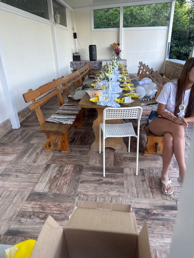 The image shows an outdoor dining area, likely a porch or patio, set up for a gathering or event. A long wooden table is laden with place settings, each featuring yellow plates and white decorations. Several white balloons are clustered near the end of the table. Wooden benches with colorful woven mats line one side of the table. A white plastic chair is positioned in front of the table. A woman in a white t-shirt and denim shorts is seated at the far end of the table, holding a decorative stick. In the foreground, a cardboard box is visible. The background features a wall, large windows overlooking greenery, and an outdoor speaker system. The floor is tiled with a wood-like pattern. The location is identified as Varbak, Bulgaria.