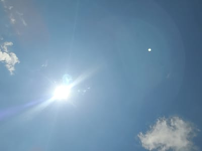 The image displays a bright, clear blue sky during daytime in Gromshin, Bulgaria. The sun shines intensely from the upper right quadrant, creating prominent lens flare effects, including radial light rays and a distinctive blue-purple streak. A secondary, circular green and white lens flare is visible in the lower-middle left of the frame, appearing as a reflection of the sun's brightness. Several white, fluffy cumulus clouds are scattered across the sky; a larger formation is in the upper left corner, and smaller clouds are seen towards the lower right. The photograph is taken looking upwards towards the expansive sky, highlighting the vibrant blue and white hues under sunny conditions. No people, specific objects other than the sky elements, or text are visible.