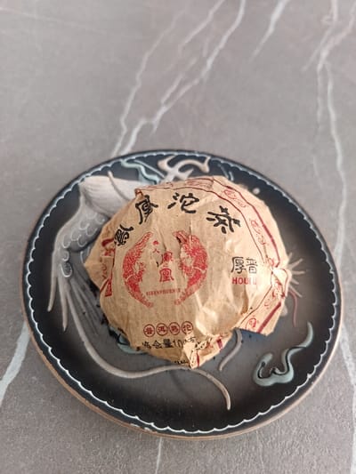 A wrapped cake of tea rests on a decorative plate. The plate has a dark gray background with a light gray raised dragon design and a tan edge. The tea is wrapped in light brown paper with red Chinese characters and a phoenix logo, labeled "VIRENPHOENIX" and "HOUUU." The paper is creased and folded around the cake. The background is a gray surface with lighter gray veining.