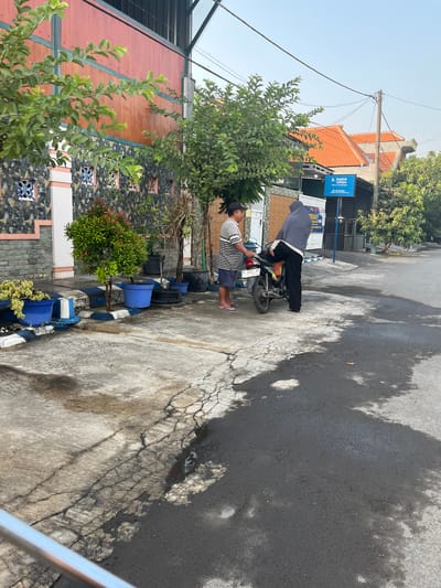 In Kedungkendo, Indonesia, a paved street with patches of cracked concrete and dark asphalt is the setting. Two individuals are near a motorcycle. One person, a man wearing a striped t-shirt and shorts, stands on the left. The other person, a woman wearing a grey hijab and black pants, is leaning over the motorcycle, with her right leg bent over the seat. The motorcycle is black and silver with a license plate that reads "W 4322 RB".
To the left of the scene, a building with a red wooden facade and a stone-tiled lower portion is visible. Several potted plants, including a small tree with red-tipped leaves in a blue pot, are arranged along the building's edge. Behind the motorcycle, a blue sign with white lettering reads "English Centre: Centre of Learning English". In the background, orange-tiled roofs of houses and lush green trees complete the environment. Power lines are strung across the sky, and a concrete utility pole stands on the right.