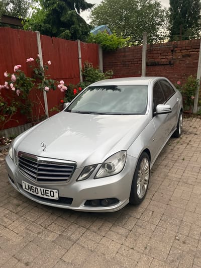 A silver Mercedes-Benz E-Class saloon, bearing the license plate LN60 UEO, is parked on a paved driveway. The car is positioned at an angle, facing slightly towards the viewer's right, with its front end clearly visible. Behind the car, a weathered red wooden fence runs horizontally across the image, with concrete posts interspersed along its length. Rose bushes with pink and red blooms are climbing the fence on the left side of the frame. To the right of the wooden fence, a section of red brick wall is visible, some bricks missing from its upper section. Overgrown green trees and foliage are visible beyond the fences and walls, suggesting a residential or semi-rural setting. The sky is overcast. The image appears to have been taken outdoors during daylight hours. The scene is located in the city of Wolverhampton, United Kingdom, with the registration plate indicating the vehicle was registered in the UK in September 2010.
