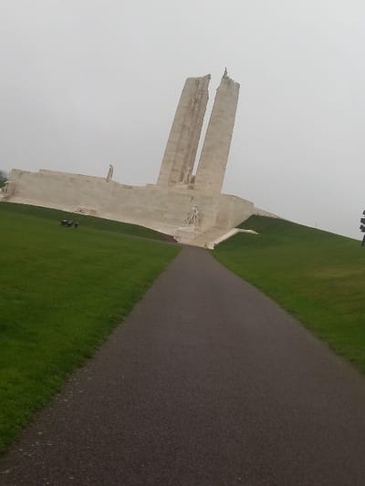 A stone memorial stands next to a green grassy field. A pathway bisects the field, leading directly to the monument. The sky is overcast, casting a muted light. The monument appears aged and worn, with visible architectural details. Its shape suggests a commemorative structure, possibly related to a war or significant historical event.