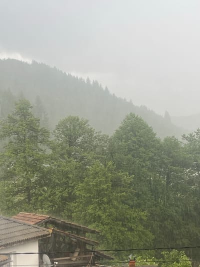 The image depicts a heavy rain shower over a forested, mountainous landscape and a residential area in Elhovets, Bulgaria.

In the foreground, occupying the lower left quadrant, is the upper portion of a residential building. It features a white wall and two distinct roof sections: one with dark gray corrugated tiles and another with terracotta-colored flat tiles. A white satellite dish is mounted on the white wall. To the right of the main roof, a wooden structure, possibly a balcony or a pergola, is visible, with its wooden beams and roof appearing wet. Several dark, horizontal power lines or cables traverse the lower-middle section of the image, running above the building. On the far bottom right, a terracotta-colored chimney pot and some light green foliage are partially visible.

The midground is dominated by a dense canopy of mature deciduous trees with vibrant green leaves, which appear wet and reflective from the rainfall. Numerous vertical streaks of rain are clearly visible against the foliage.

In the background, a series of forested mountains or hills extends across the upper half of the image. These distant tree lines appear as subdued, dark green silhouettes, significantly obscured by the heavy rain and atmospheric haze, which reduces visibility. The sky above is entirely overcast, presenting as a uniform light gray or white, indicating dense cloud cover associated with the ongoing precipitation. No individuals or animals are visible within the frame.