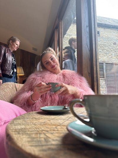 This image captures an indoor scene in Kilmersdon, United Kingdom, featuring four individuals within a warm, rustic environment that looks out onto an older stone building.
In the foreground, a light-skinned woman with curly, light brown hair styled in a high bun smiles directly at the viewer. She is seated in what appears to be an armchair, wearing a voluminous, textured pink faux fur top and a bright pink garment on her lap. Her left wrist displays a metallic, possibly silver-toned, coiled bracelet. She holds a light teal-colored ceramic cup with both hands, its rim resting just below her chin. On the wooden table in front of her, which exhibits a distinct grain and carved or etched patterns, sits another matching light teal ceramic saucer with a small spoon, and partially visible in the extreme lower right foreground, a second matching cup.
In the mid-ground, to the left of the smiling woman, a second light-skinned woman with blonde hair stands, looking downwards and to her left. She wears a dark red or maroon leather jacket over a dark top with a graphic design, and blue jeans. Behind her, a wooden-paneled wall and a lamp with a patterned shade are visible.
The large window to the right of the central woman offers a view of the exterior. Through the window, the upper torsos of two more individuals are partially visible. One appears to be a male youth with light brown hair, wearing a dark hooded top, looking away to the right. A second, younger individual is partially obscured by him and the window frame, wearing a light-colored top. Beyond the window, a substantial building constructed from light-colored, coursed stone blocks is visible, characteristic of traditional British architecture, with a visible roofline and a rectangular window opening with horizontal slats below. The exterior lighting suggests daytime. The overall interior environment is softly lit, with warm tones from the wood and fabric, contrasting with the brighter, stone exterior.