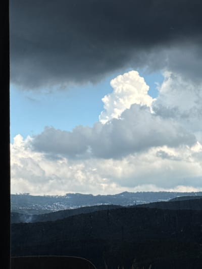 A photograph captures a panoramic view of the city of Đa Lộc, Vietnam, dominated by dramatic cloud formations and mountainous terrain. The foreground is defined by the dark, silhouetted forms of rolling hills, their dense foliage obscuring any ground-level details. These hills create a stark contrast against the sky, which is filled with a dynamic interplay of light and shadow.

The sky is a prominent feature, with vast expanses of both dark, ominous storm clouds and bright, fluffy cumulus clouds. The storm clouds, particularly in the upper left quadrant, appear heavy and laden, suggesting an approaching or receding weather system. In stark contrast, a large, cauliflower-like white cloud formation stands out against a patch of bright blue sky in the upper center of the image. This bright cloud is illuminated from above, indicating sunlight breaking through or a gap in the darker cloud cover. Below these, a layer of lighter, scattered clouds stretches across the horizon.

In the mid-ground, a town or city is visible nestled within the undulating landscape. Numerous buildings, appearing as a cluster of small structures, are scattered across a valley or plateau. The density of these buildings suggests a populated area. A faint wisp of smoke or steam rises from one of the darker hills in the mid-ground, possibly indicating localized activity or atmospheric conditions.

The overall impression is one of natural grandeur, with the weather and landscape taking center stage. The absence of any visible people, vehicles, or specific man-made structures in the immediate foreground or mid-ground, beyond the distant settlement, keeps the focus firmly on the environment. The image is shot from an elevated perspective, looking out over the landscape and towards the horizon, with a dark, vertical element on the left edge of the frame suggesting it was taken from within a structure or through a narrow opening.