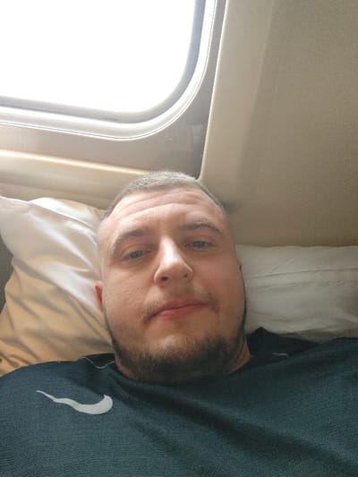 A male individual, estimated to be in his 20s to 30s, is captured in a self-portrait within an enclosed compartment, located in Druzhinino, Russia. He possesses short, dark hair, light-colored eyes, and a short beard and mustache, presenting a neutral expression while looking directly into the camera. He wears a dark-colored t-shirt with a light-colored Nike 'swoosh' logo visible on the left chest. The individual is reclined, supported by a white pillow. To his upper left, a rounded rectangular window displays an overexposed, bright white exterior, suggesting daylight. The interior paneling of the compartment surrounding the window and to the right is light beige or cream in color.