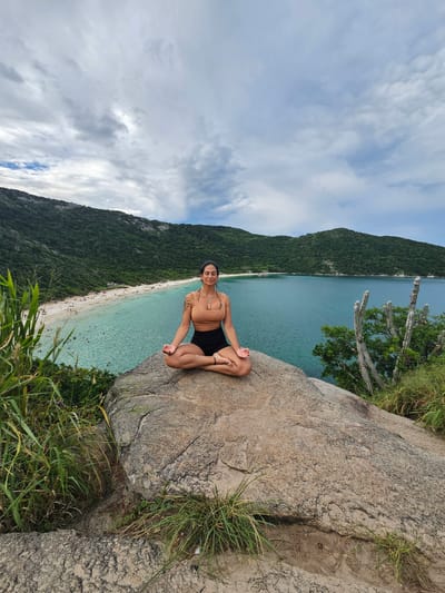 The image depicts a female in her 20s or 30s seated in a lotus position (Padmasana) on a large, textured gray-brown rock outcrop in Arraial do Cabo, Brazil. She is positioned in the immediate foreground, looking directly at the camera with a slight smile. She wears a light brown sports bra and black shorts, with a light-colored pendant visible on her chest and tattoos on her left arm. Her hands rest on her knees, fingers forming a mudra.
The scene overlooks a scenic bay. In the midground, a crescent-shaped white-sand beach (Praia do Forno) is visible along the curve of the bay, populated by numerous small figures of individuals in the sand and clear, turquoise-blue water, indicating recreational activity. The bay's water transitions to a deeper blue further from the shore. Beyond the beach, densely vegetated green hills rise steeply, forming the natural backdrop of the bay.
To the immediate left and right of the subject and rock, green grasses and other foliage are present. On the right side, several tall, columnar cactus-like plants with a weathered appearance are visible among the green vegetation. The sky above is partly cloudy, displaying a mix of light and dark gray clouds with patches of blue. The overall lighting is diffused.