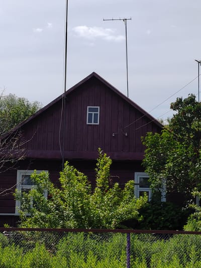 This media file is a photograph of a dark red wooden house with a steep gabled roof. The house has at least two visible windows, both with white frames. One is a small, square window located in the upper gable, and another is a larger, rectangular window visible on the lower level. The house is surrounded by lush green trees and foliage, with a chain-link fence in the foreground. Above the house, two tall antenna poles are visible, one with a horizontal antenna structure at its top. The sky is overcast with some scattered clouds. The overall scene suggests a rural setting, possibly a residential house in the countryside. There is no visible text in the image. The lighting indicates it is daytime, likely during daylight hours, under cloudy or partly cloudy weather conditions. The location context provided is Воўчынскі сельскі Савет, Belarus.