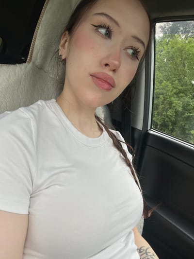 A young Caucasian woman with long brown hair in a braid, wearing a white t-shirt, is sitting in the passenger seat of a car. She is looking out the window to her right, with a slight smile. Her makeup includes winged eyeliner and pink blush. The car interior is visible, including a fluffy seat cover and the car door. Outside the window, trees are visible, suggesting a rural or suburban setting. The image appears to be taken during the daytime, under overcast skies.