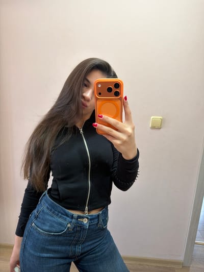 A young woman is depicted taking a mirror selfie in an indoor setting, likely in Rudartsi, Bulgaria. She holds an orange smartphone with a prominent camera array and a circular MagSafe-compatible ring in her right hand, partially obscuring her face, though her lower nose and lips are visible. She has long, dark brown hair that flows downwards. Her fingernails are painted with bright red polish. She is dressed in a black long-sleeved top with a silver zipper running down the front, paired with blue denim jeans.

The background features a plain, light-colored wall, possibly pale pink or beige, on which a gold-colored square light switch or power outlet is mounted towards the top left. The bottom of the image reveals a light-colored wooden floor separated from the wall by a white skirting board. The lighting appears to be natural, suggesting daytime. No discernible text, specific emotions, or particular weather conditions are evident in the image.