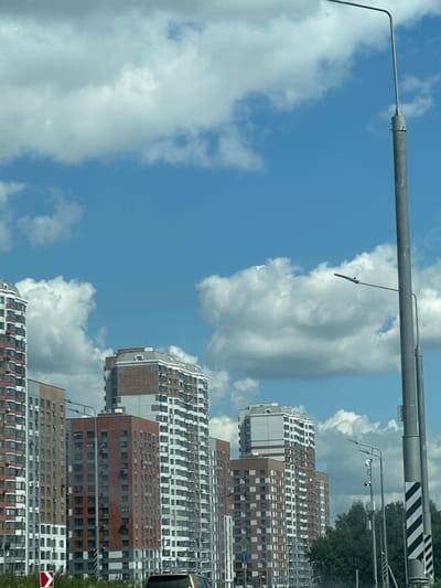 A high-angle, eye-level shot captures a modern residential area in Butovo, Russia, under a bright, cloudy sky. Several tall apartment buildings, characterized by their red and white facades with numerous windows, dominate the frame. In the foreground, a tall, grey lamppost with two lights stands prominently on the right side of the image. Another lamppost, further back and to the left, also features a single light. The buildings are situated in a dense urban setting, with a line of trees visible behind them on the right. A red and white directional traffic sign is present in the lower left corner, suggesting the presence of a roadway. No people are visible in the scene.