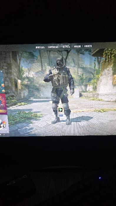 A vertically oriented image shows a video game displayed on a screen. The game features a soldier character standing in a paved outdoor environment with some greenery. The menu options on the left are written in Russian. Below the game screen, icons for various programs are visible. The rest of the image is dominated by a dark background including a computer keyboard and mouse on the right.