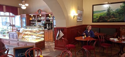 This image depicts the interior of a cafe, likely located in the city of Viljandi, Estonia, given its ambiance and style. The cafe features a warm and inviting atmosphere, characterized by its wooden furniture, dim lighting, and traditional decor.
In the foreground, several red velvet chairs with wooden frames are arranged around small, round tables. One table in the mid-ground has a woman seated at it, looking towards the right. She is wearing a blue jacket and holding a dark object in her hand. A plate with a cup of coffee and some pastries is visible on the table.
To the left, a display case showcases a variety of pastries and desserts. Behind it, a counter area is stocked with bottles of alcohol and glassware, suggesting a bar or service area. Further back, coffee machines and other kitchen equipment are visible.
The walls are decorated with patterned wallpaper and framed artwork, including a large landscape painting above the seating area where the woman is sitting. A chandelier hangs from the ceiling, providing soft illumination. Newspapers are neatly displayed on a rack attached to a red wooden panel. The flooring consists of a tiled pattern in shades of grey and white. The overall impression is of a well-established, possibly historic, cafe with a cozy and classic aesthetic.