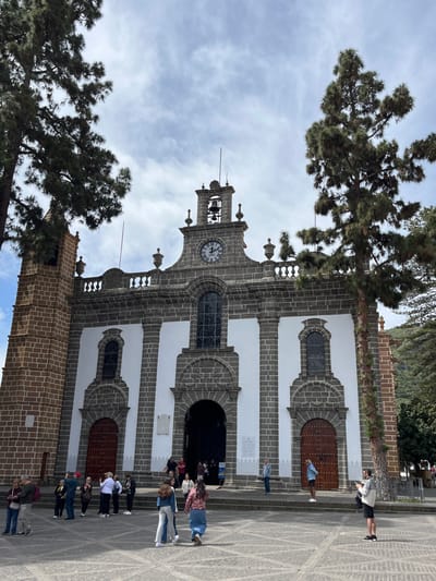 The image shows the Basilica de Nuestra Señora de la Candelaria in Teror, Gran Canaria, Spain. The basilica is constructed of grey stone with white accents. It features two side doors and a large entrance, all with wooden doors. There is a clock face on the facade above the entrance and a bell tower. There are two trees on the left side of the image. There are several people standing in front of the building, some looking at their cell phones and others interacting with each other. The ground is made of grey cobblestones.