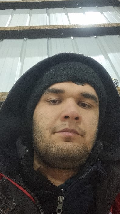 A man with dark hair and stubble wears a black hooded jacket indoors. He looks directly at the camera, his expression neutral. He also wears a black beanie. The background reveals a wall of white panels secured with wooden beams. The scene is brightly lit.
