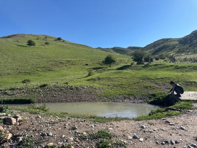 The image depicts a natural outdoor scene with rolling green hills under a clear blue sky, likely in the vicinity of Kashka-Suu, Kyrgyzstan. In the foreground, a small, shallow body of water, possibly a stream or pond, is visible. The water appears murky and reflects the surrounding landscape. The bank of the water is lined with rocks and sparse vegetation.

To the right of the water, a young person is crouched on a concrete-like object, holding a stick and seemingly interacting with the water. This individual is wearing dark clothing with white accents and a backpack. The surrounding terrain is uneven and covered with grass and rocks. In the background, the grassy hills are dotted with a few scattered trees and some patches of lighter-colored vegetation, possibly dry grass or flowers. Further in the distance, more hills form the horizon, and a rustic wooden fence is visible on the right side of the frame. The overall impression is one of a tranquil, rural landscape on a sunny day.