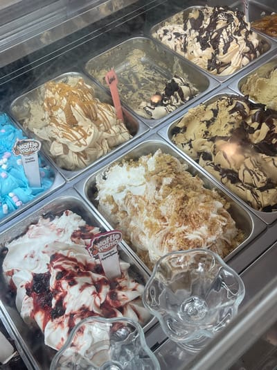 The image depicts a refrigerated display case filled with various flavors of ice cream. The location is identified as Nea Makri Municipal Unit, Greece. The ice cream is arranged in rectangular metal containers.
Flavors visible include:
* A light-colored ice cream with dark chocolate drizzle
* A light brown ice cream with chocolate pieces
* A white ice cream with a sprinkled, textured topping
* A light brown ice cream with a drizzle, labeled "Lotus"
* A blue ice cream with marshmallows
* A white ice cream with dark red swirls.
Two empty glass sundae dishes are placed near the red swirled ice cream. Waffle Mania labels are present on the blue and red swirled ice creams.