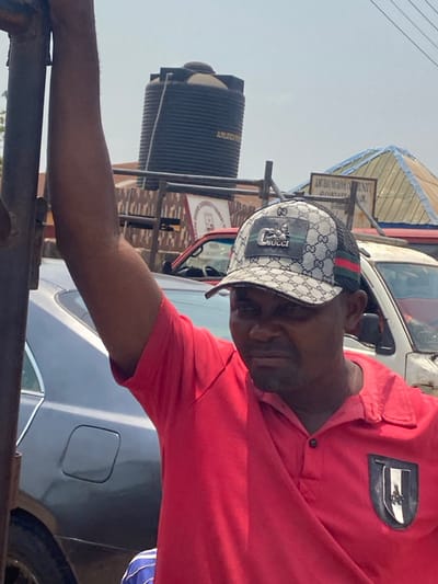 An adult male is partially visible in the foreground, wearing a red polo shirt with a dark emblem on the left chest and a white and grey patterned trucker cap featuring a "GUCCI" logo and green/red stripe detail. His right arm is raised, grasping a vertical metallic pole or frame to his left. He has a short beard or stubble and a neutral expression, looking slightly downwards.
In the mid-ground, a dark grey sedan is parked to the left, partially obscured by the man and the pole. To the right of the man, a white vehicle (possibly a van) is parked. Further back, between these vehicles, a red vehicle is visible.
The background features two large, black, ribbed plastic water tanks, one bearing the text "AMURUMU" or similar. Behind the tanks and vehicles, a wooden sign clearly reads "AMURO MGBOM COMMUNITY PRIMARY SCHOOL," establishing the location in Afikpo, Nigeria, near this institution. A light-colored, possibly corrugated metal, pyramidal roof is also visible behind the sign. The sky is clear, indicating daytime.